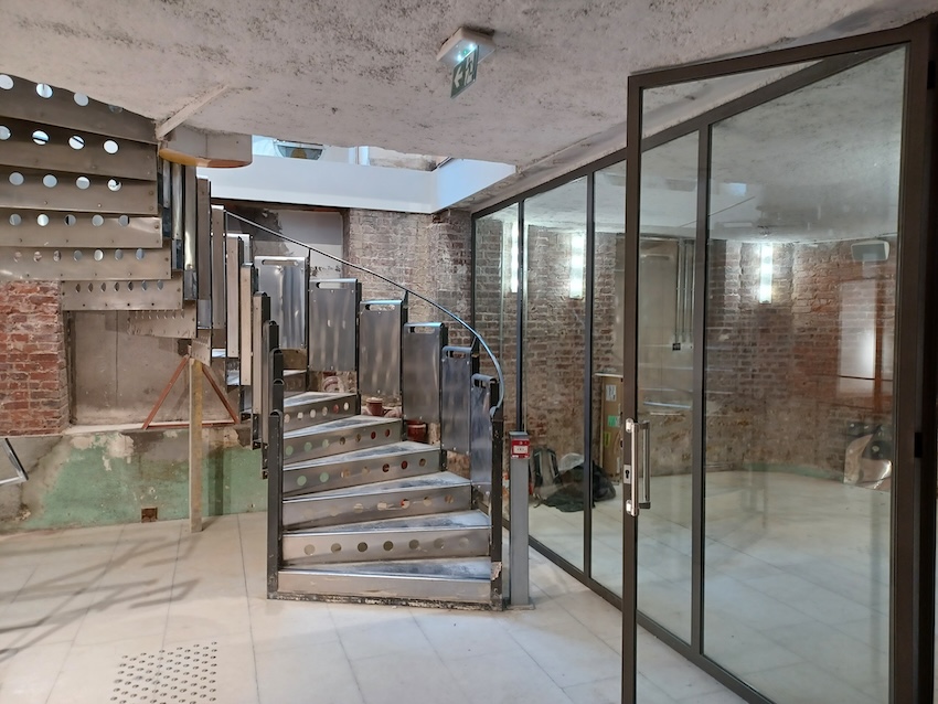 Lounge area with brick walls, glazed partition doors, and a metal staircase with perforated steps.