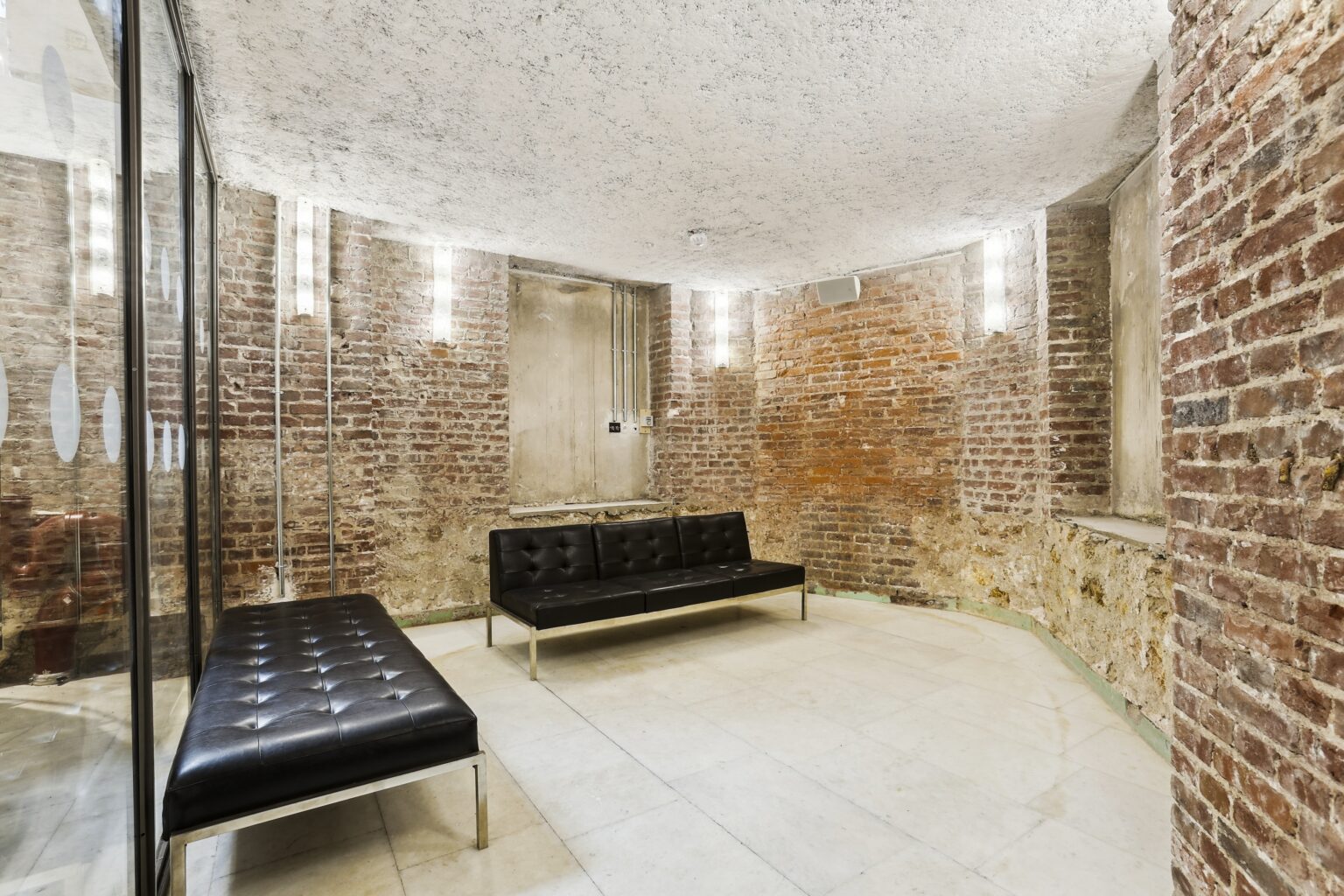 Brick-walled alcove with black seating benches and vertical wall lights, adjacent to a glass partition.