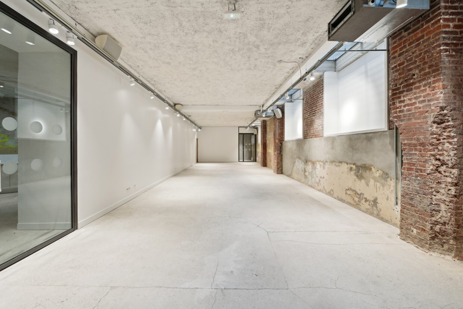 Long interior view with a white display wall and track lighting on the left, brick and frosted windows on the right.