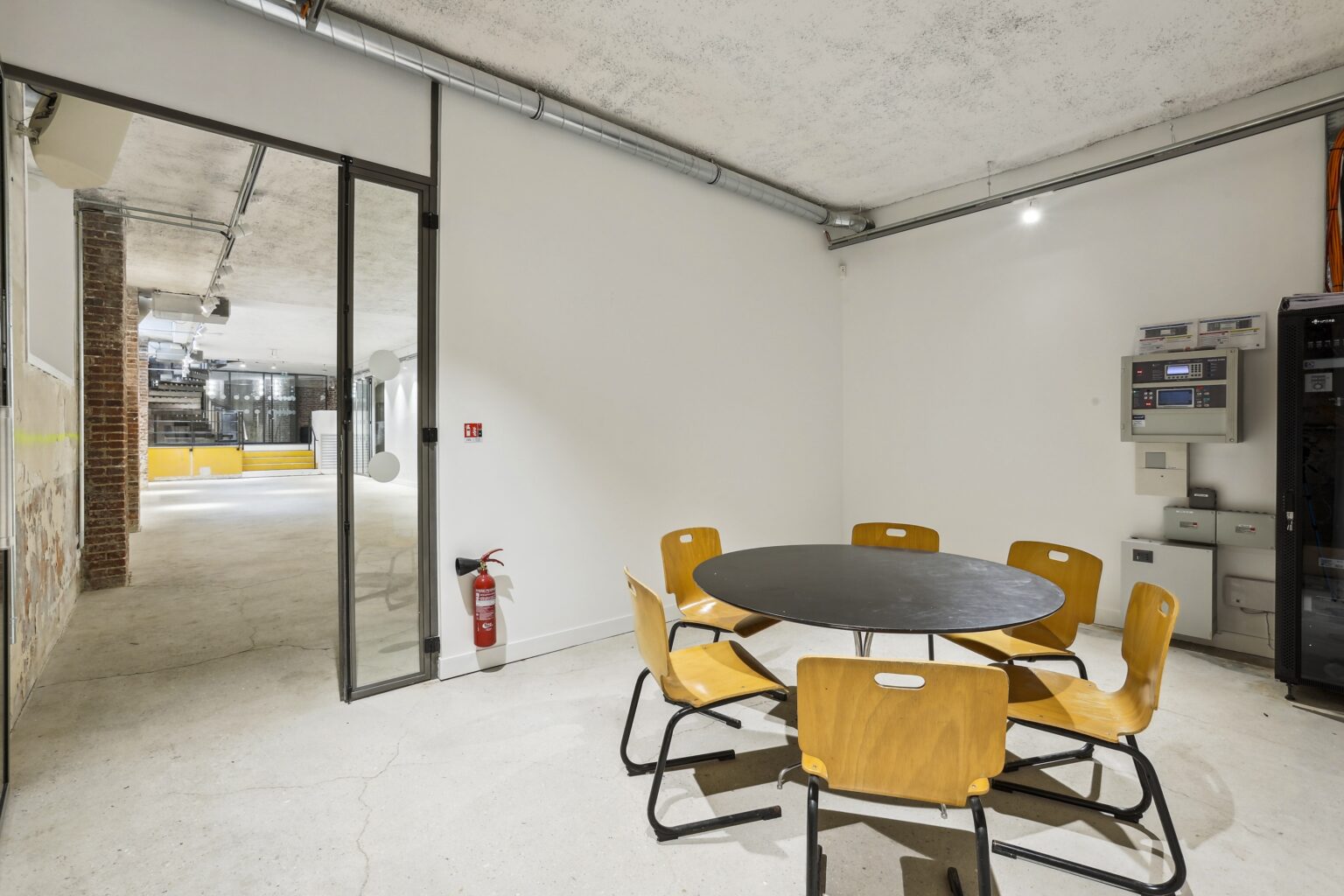 Meeting area with a round table, wooden chairs, glass door, and white walls with exposed ducting.