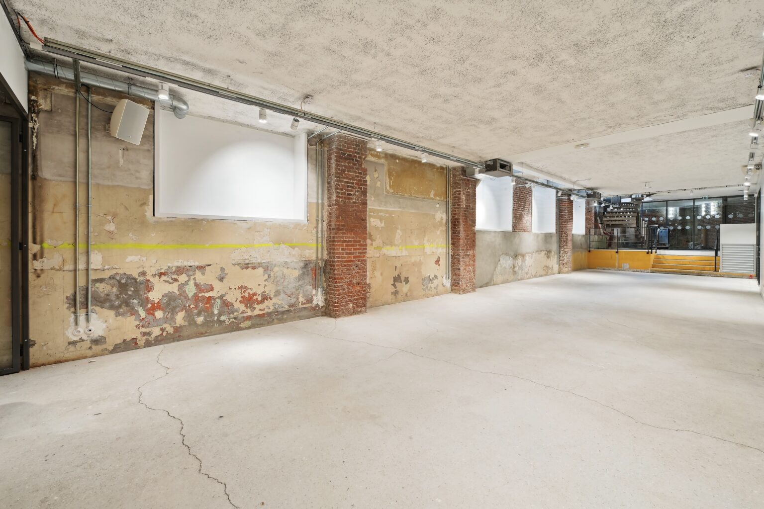 Wide interior view with distressed wall, projection screens, brick columns, and an open concrete floor leading to steps.