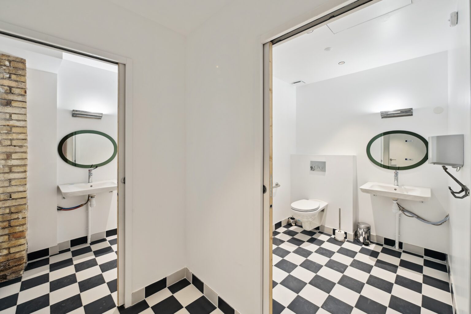 Restroom area with black-and-white checkered floor, white sinks, oval mirrors, and a toilet in the background.