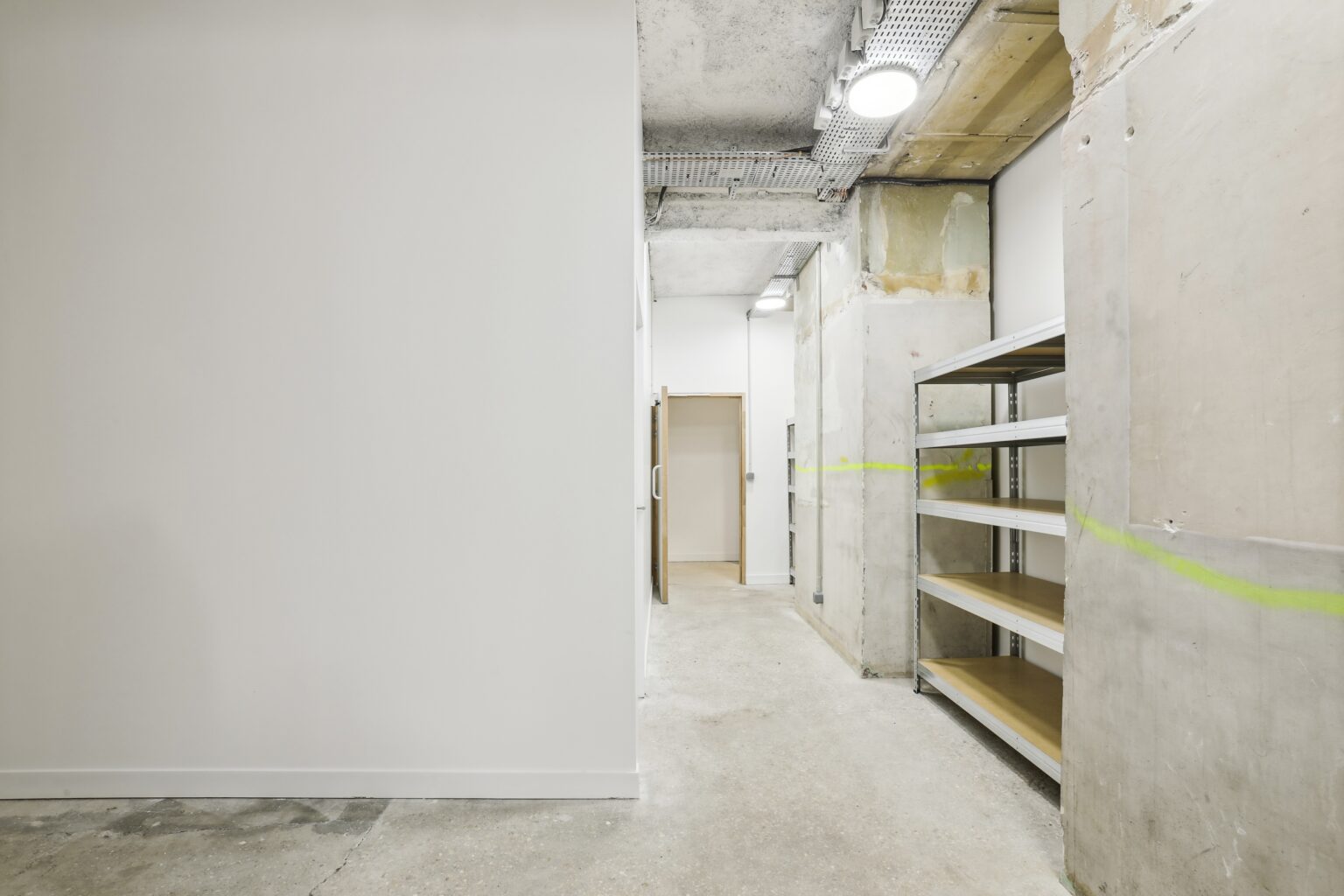 Service corridor with built-in shelving, raw concrete surfaces, and overhead lighting leading to a door.
