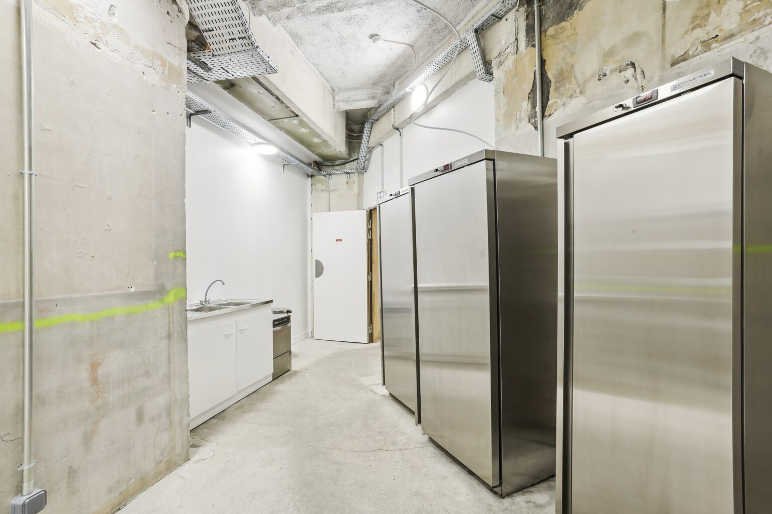 Service area with a sink unit and multiple stainless steel refrigerators under exposed ceiling cable trays.
