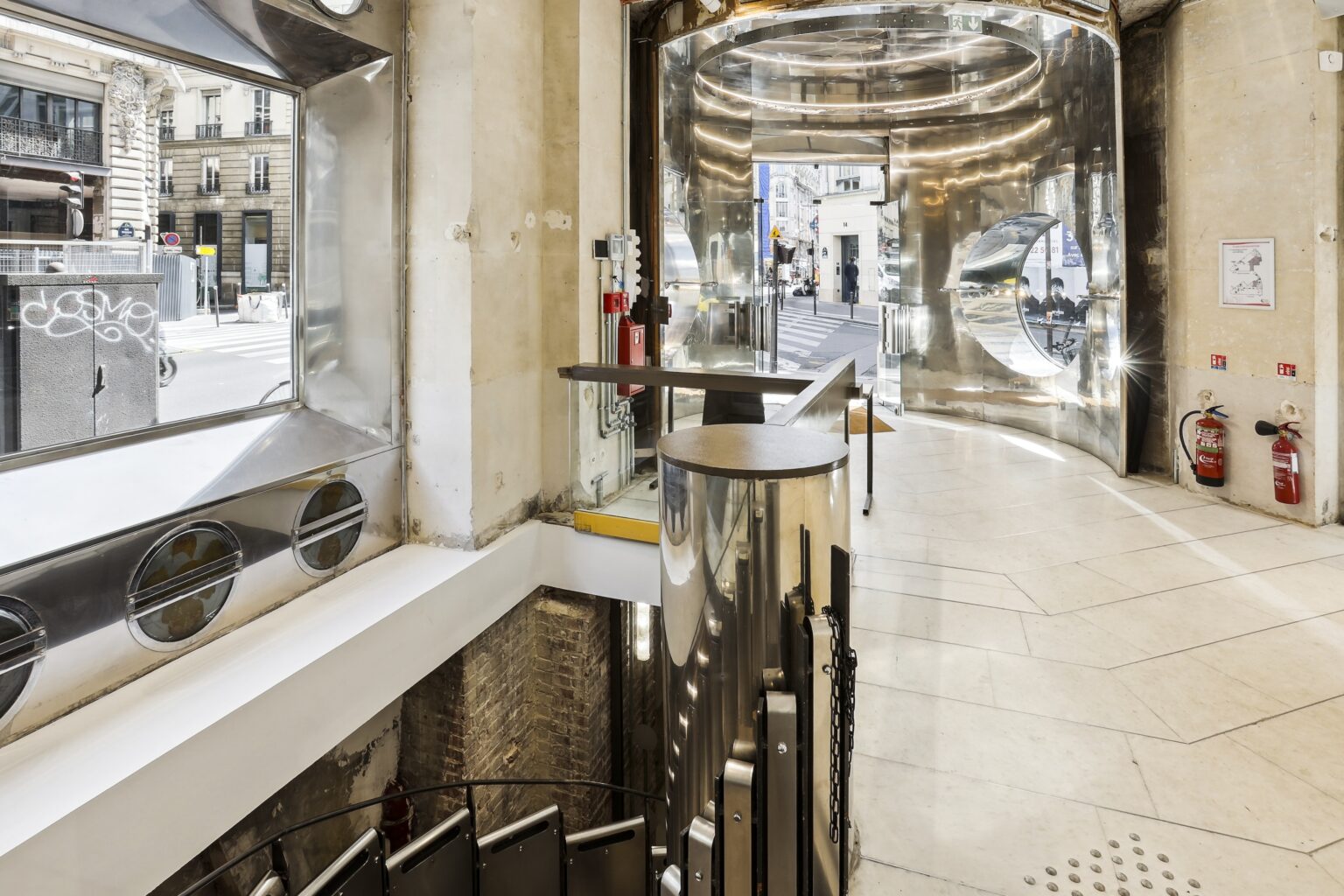 Chrome entrance portal with curved doorway, street view, display window and metal handrail overlooking the stairwell.