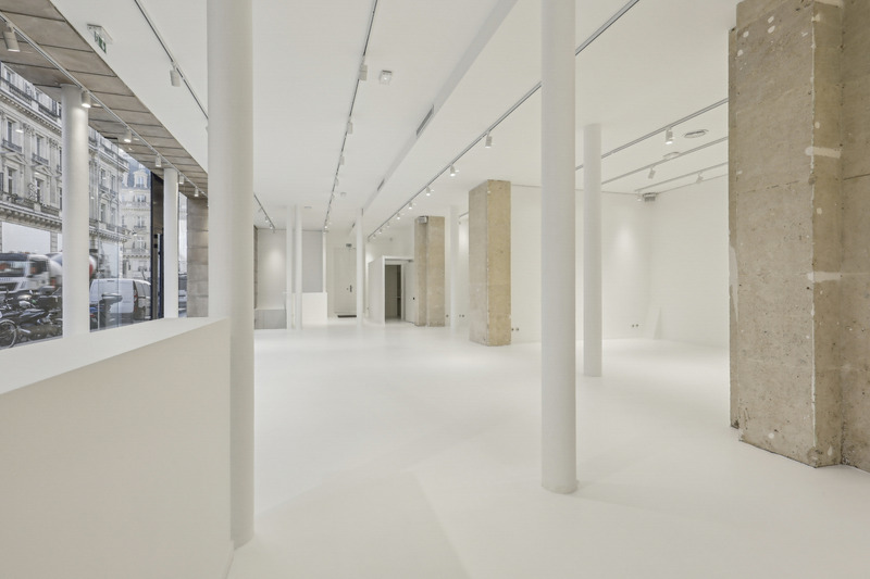 Long interior view with slim white columns, stone columns, and glass frontage on the left.