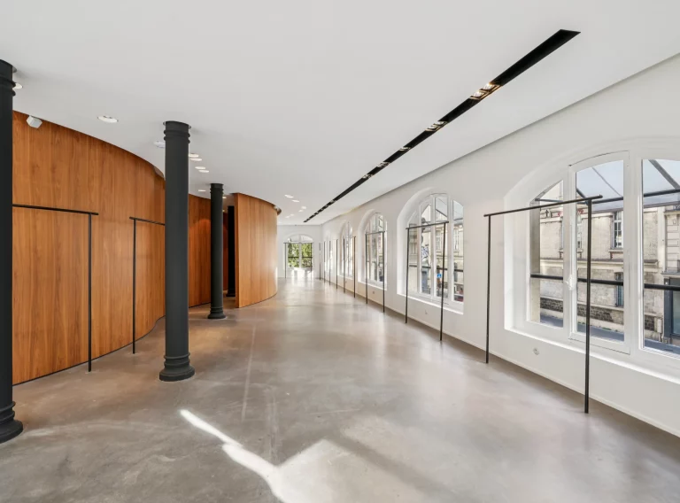 Long gallery space with arched windows facing the street and wood pod opposite.