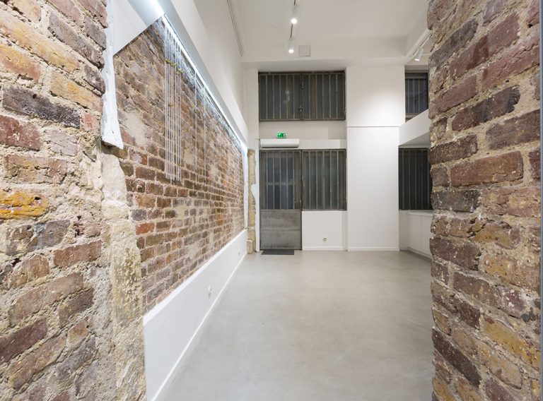 Long gallery axis with brick wall and spot lighting — Rue de la Corderie
